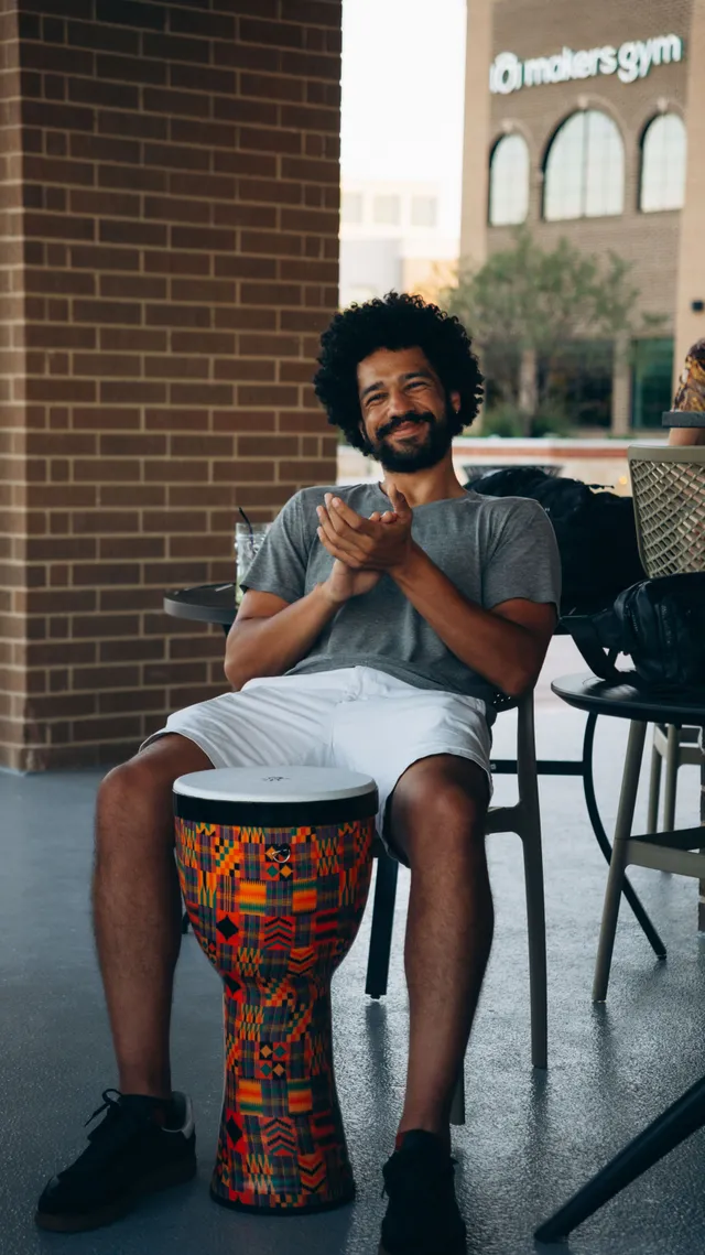 A man smiles and claps while seated with a hand drum on a patio.