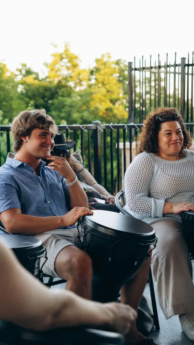 Two people play hand drums together on a covered patio.